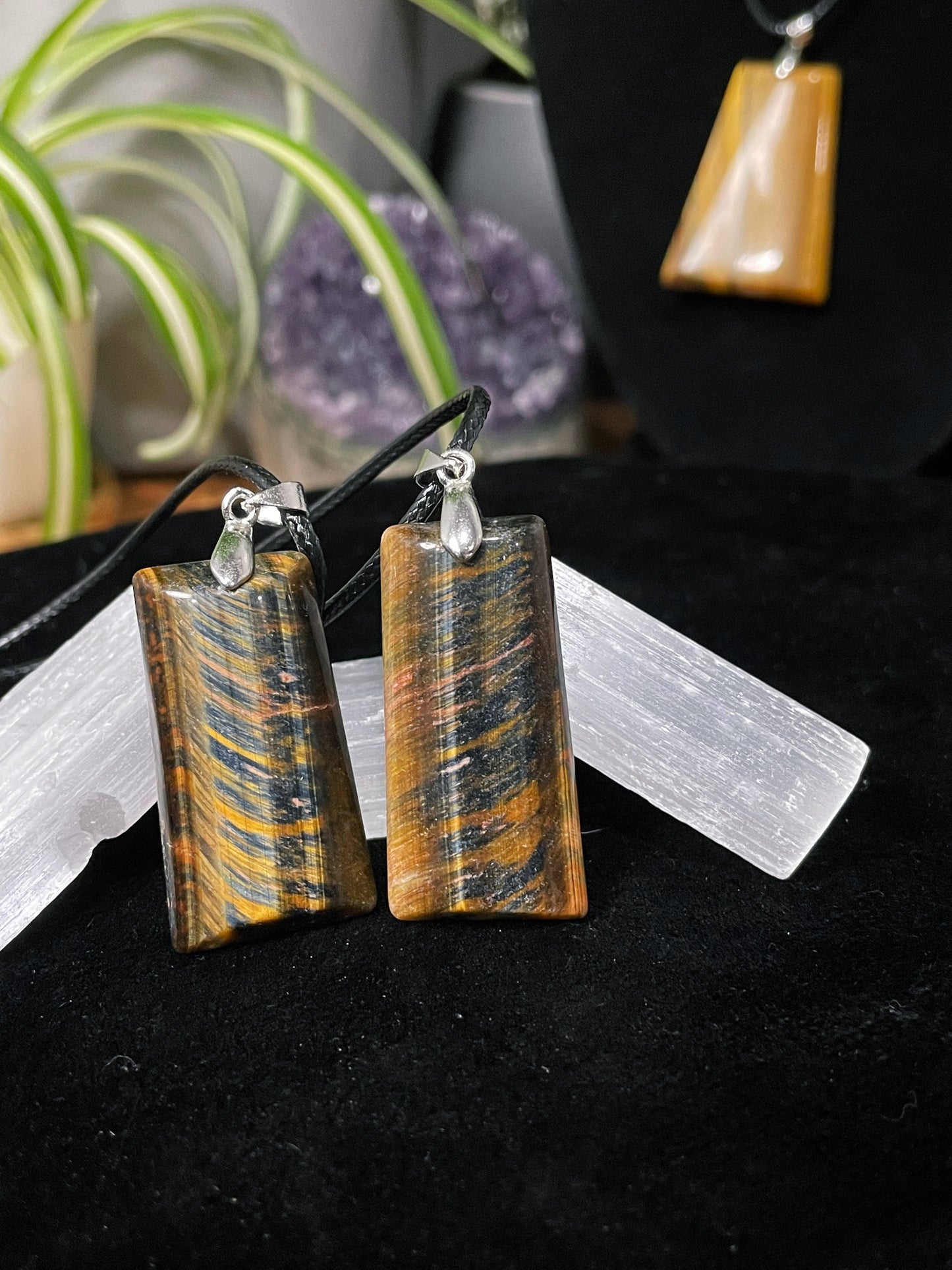 An image of a matching pair of yellow and blue tiger's eye rectangular shaped pendants on necklaces. They sit atop some selenite chunks and a black velvet surface.