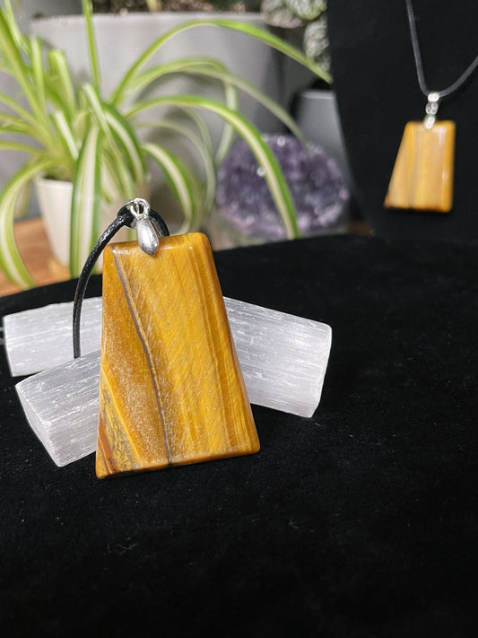 An image of a yellow tiger's eye rectangular shaped pendant on a necklace. It sits atop some selenite chunks and a black velvet surface.