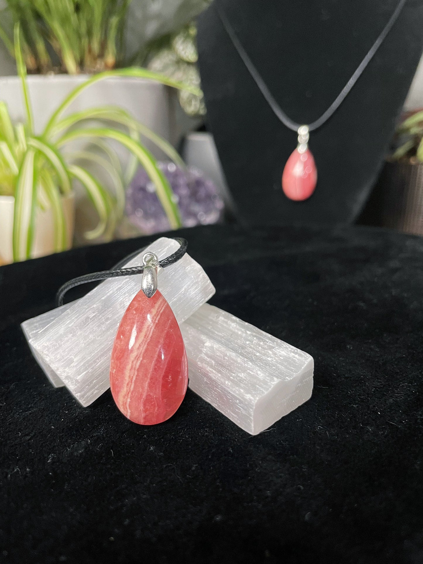 An image of a high-quality pink rhodochrosite teardrop shaped pendant on a necklace. It sits atop some selenite chunks and a black velvet surface.