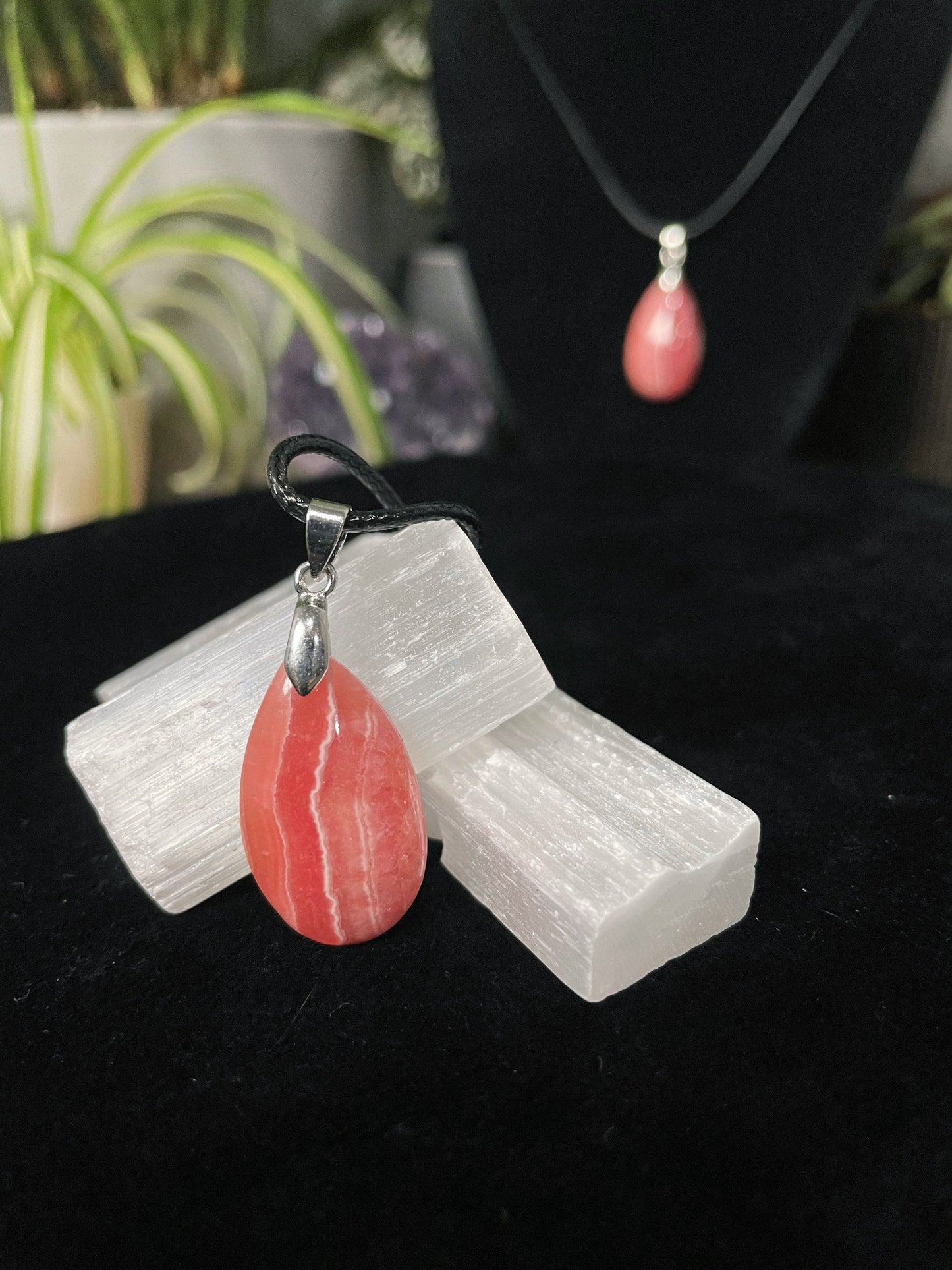 An image of a high-quality pink rhodochrosite teardrop shaped pendant on a necklace. It sits atop some selenite chunks and a black velvet surface.