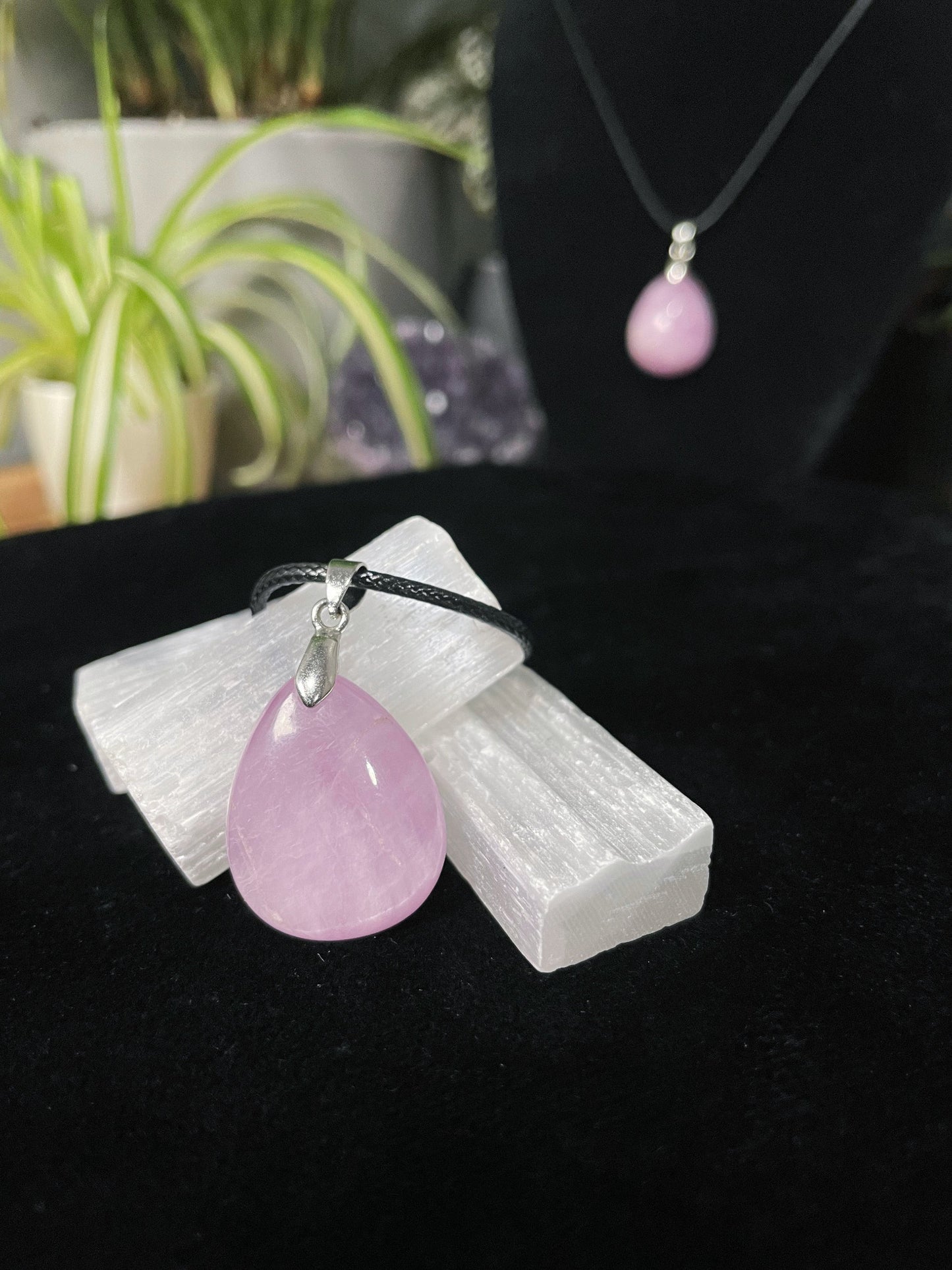 An image of a high-quality kunzite teardrop shaped pendant on a necklace. It sits atop some selenite chunks and a black velvet surface.