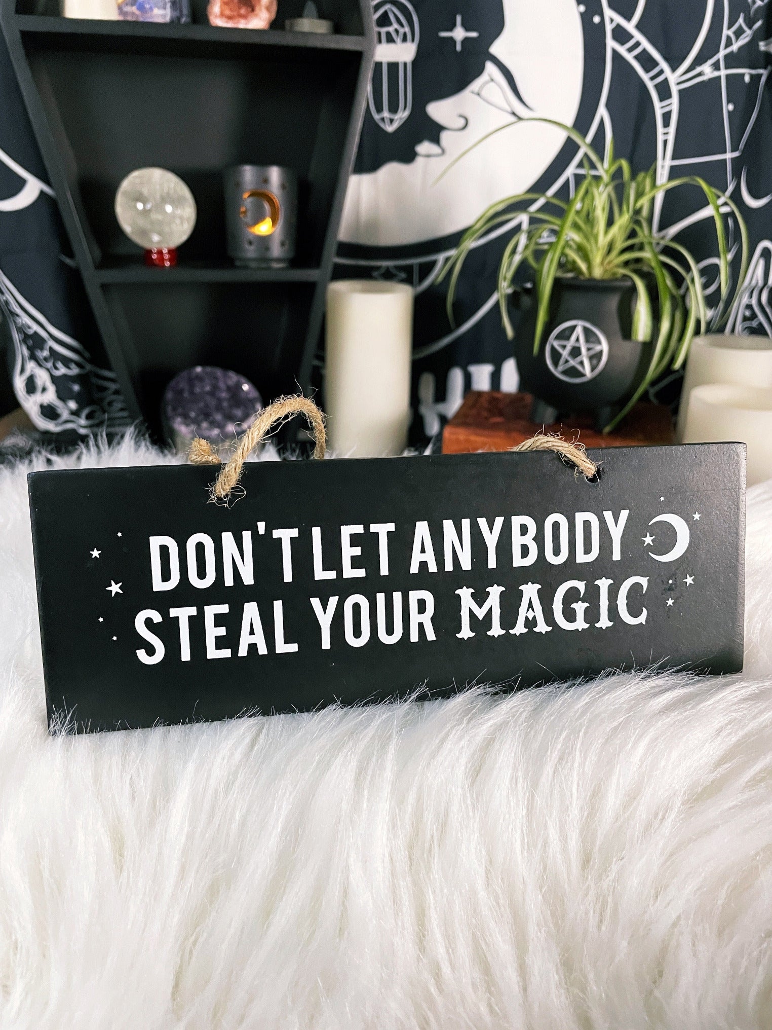 An image of a black sign with white letters that reads "Don't Let Anybody Steal Your Magic." The sign is rectangular and has a thick rope hanger attached to it, making it suitable for hanging on a wall or door. 