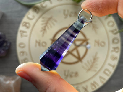 A purple and blue fluorite crystal pendulum sits atop a wooden divination board.
