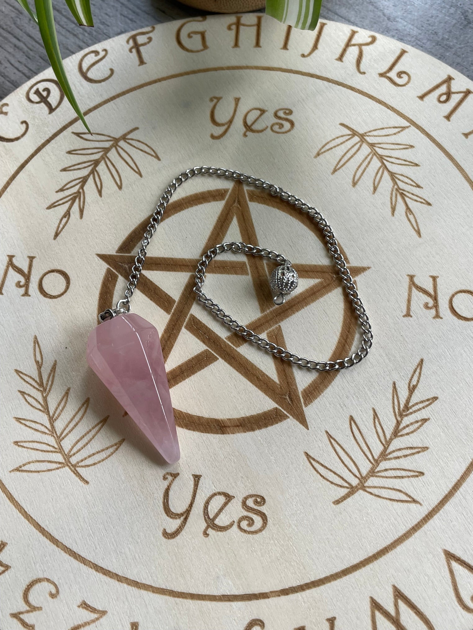 A pink rose quartz crystal pendulum sits atop a wooden divination board.