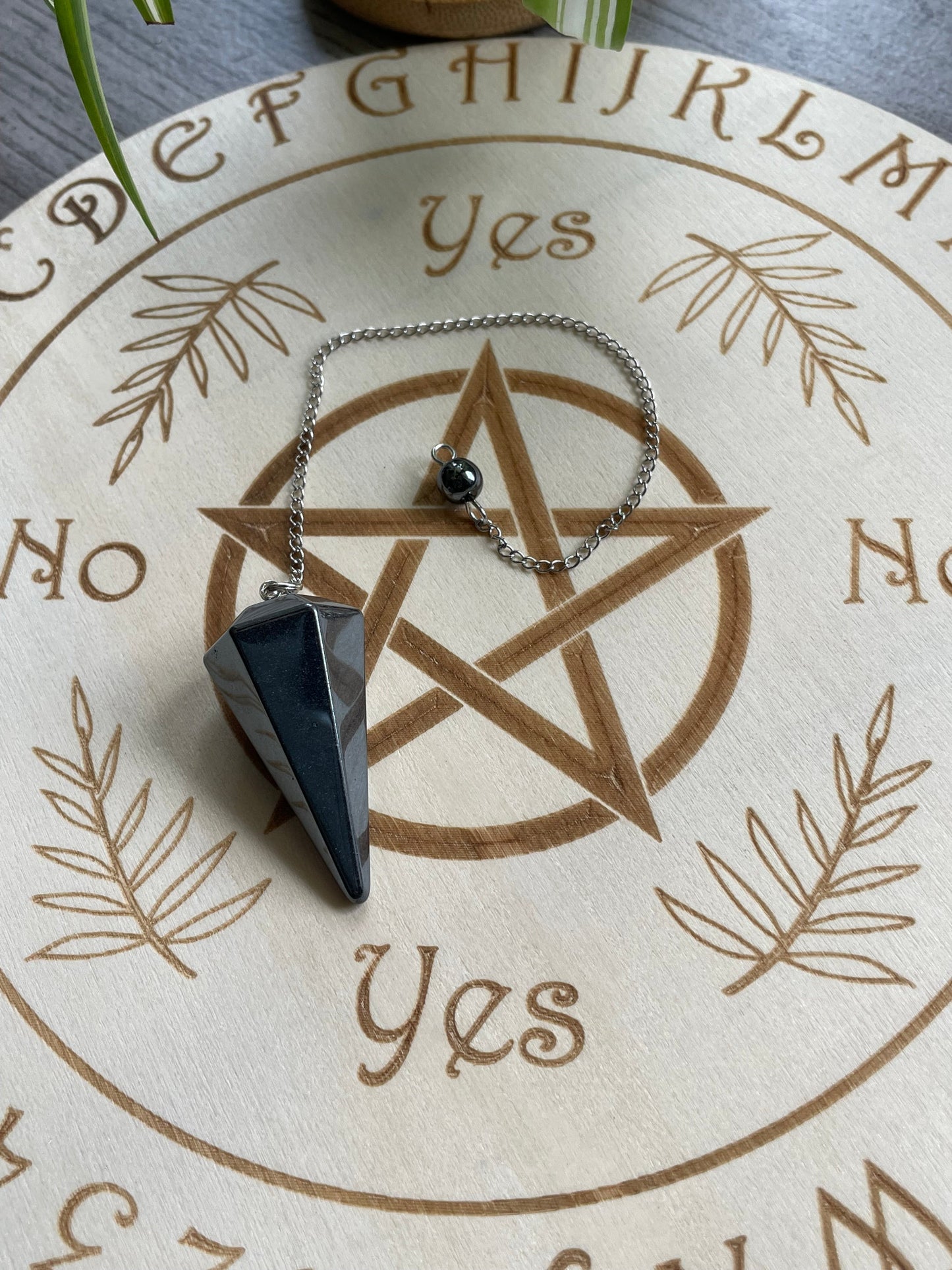 A hematite crystal pendulum sits atop a wooden divination board.