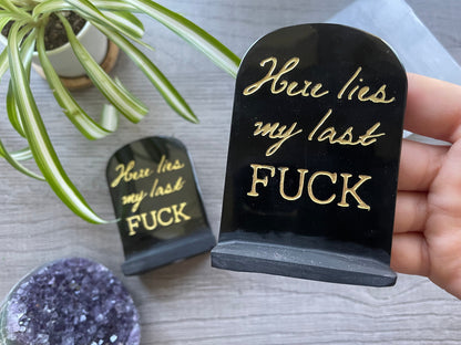 An image of a carved obsidian tombstone with the words "Here Lies My Last Fuck" written in gold letters on it. The tombstone is a humorous and cheeky statement piece that adds a touch of personality and edge to any space, and serves as a reminder to let go of negativity and embrace a more carefree and lighthearted attitude.