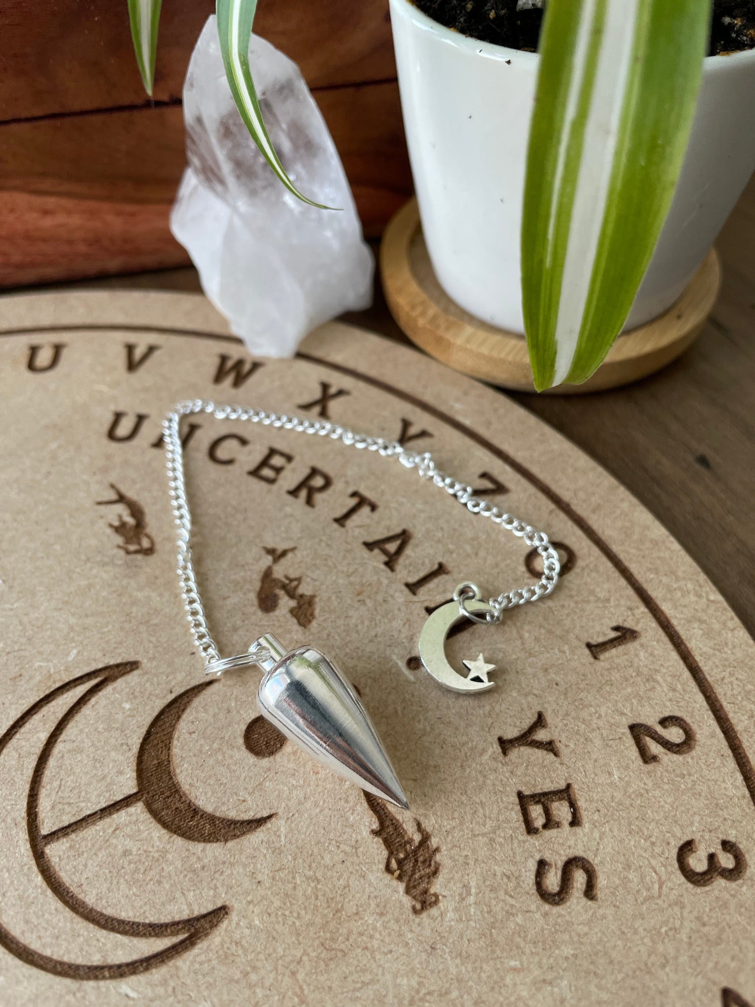 A silver coloured pendulum with a crescent moon and star sits atop a wooden divination board.