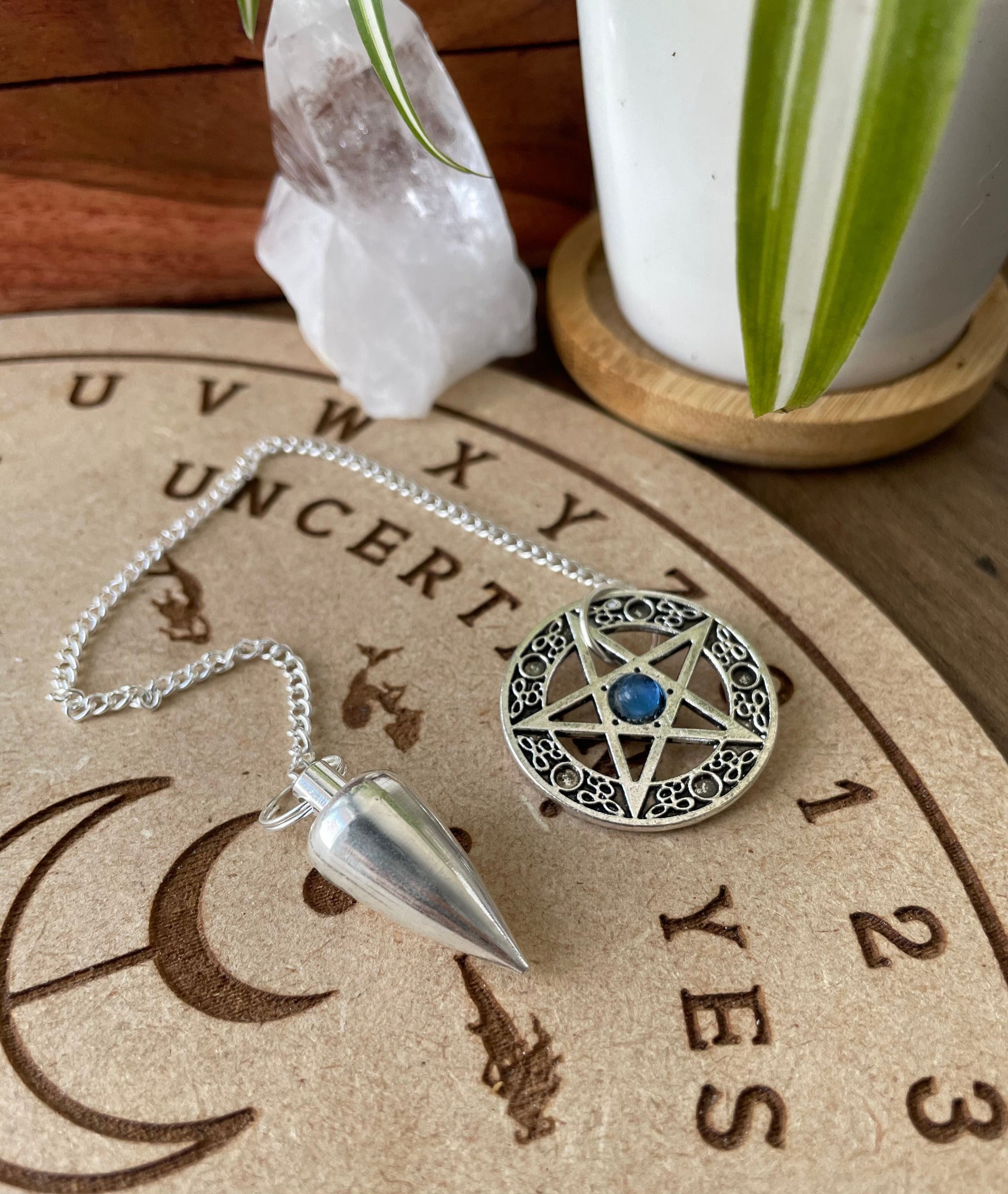A silver coloured pendulum with a pentagram sits atop a wooden divination board.