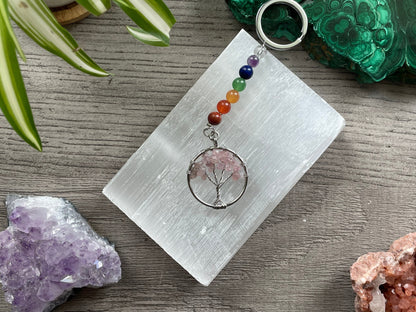 A tree of life with rose quartz crystals as the leaves on the tree is in the image. The keychain has a number of small multi-coloured beads to represent the chakras on the chain. The keychain sits atop a slab of selenite. Malachite, pink amethyst, and amethyst are nearby.