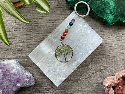 A tree of life with peridot crystals as the leaves on the tree is in the image. The keychain has a number of small multi-coloured beads to represent the chakras on the chain. The keychain sits atop a slab of selenite. Malachite, pink amethyst, and amethyst are nearby.
