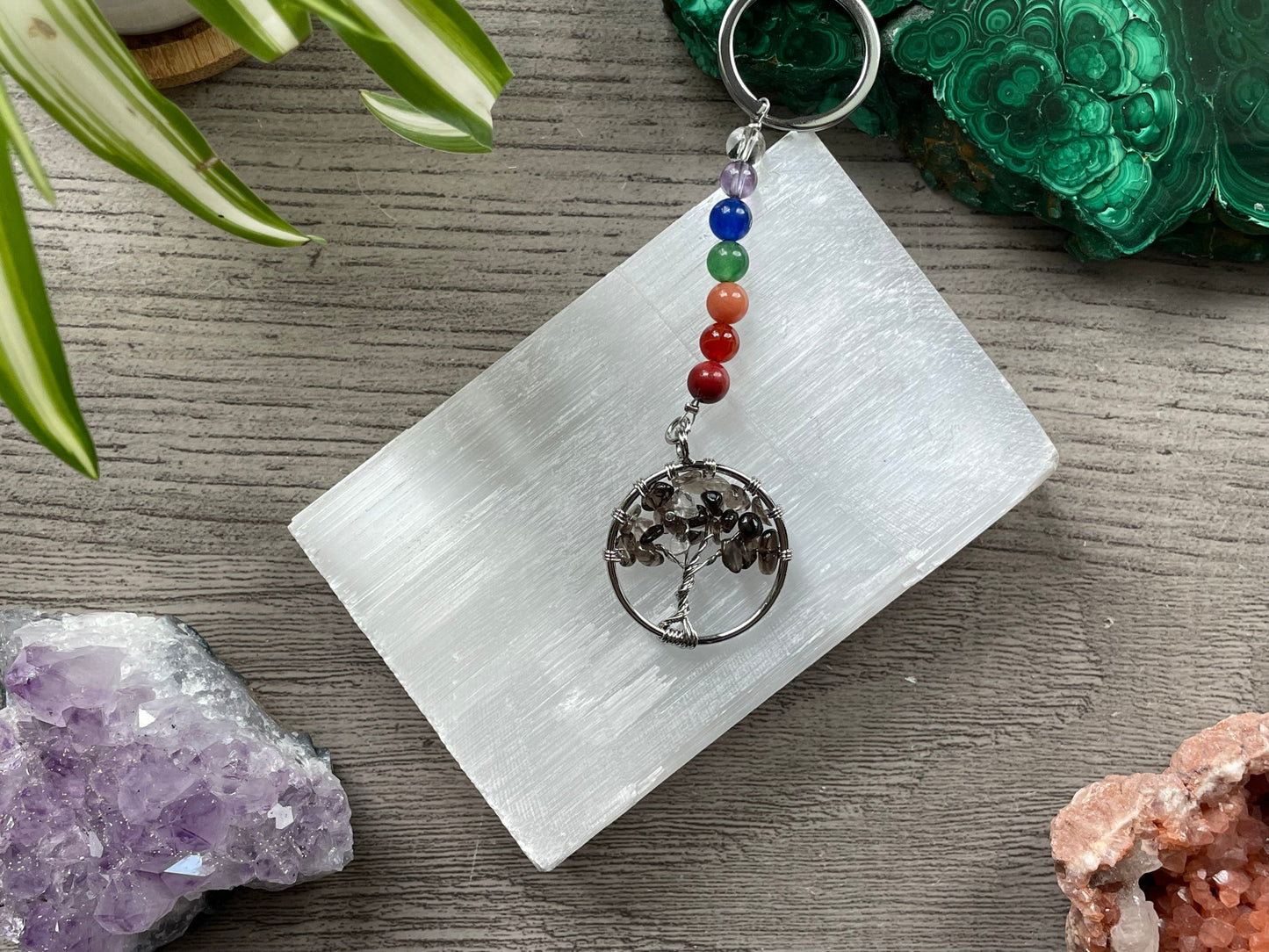 A tree of life with smoky quartz crystals as the leaves on the tree is in the image. The keychain has a number of small multi-coloured beads to represent the chakras on the chain. The keychain sits atop a slab of selenite. Malachite, pink amethyst, and amethyst are nearby.