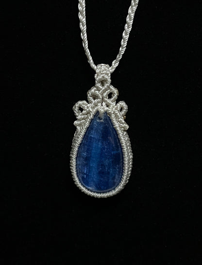 Pictured is a blue kyanite cabochon wrapped in macrame thread. A gothic book and flowers are nearby.