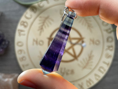 A purple and blue fluorite crystal pendulum sits atop a wooden divination board.