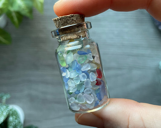 Pictured is a small glass vial with a cork stopper. Inside the jar are sea glass chips.