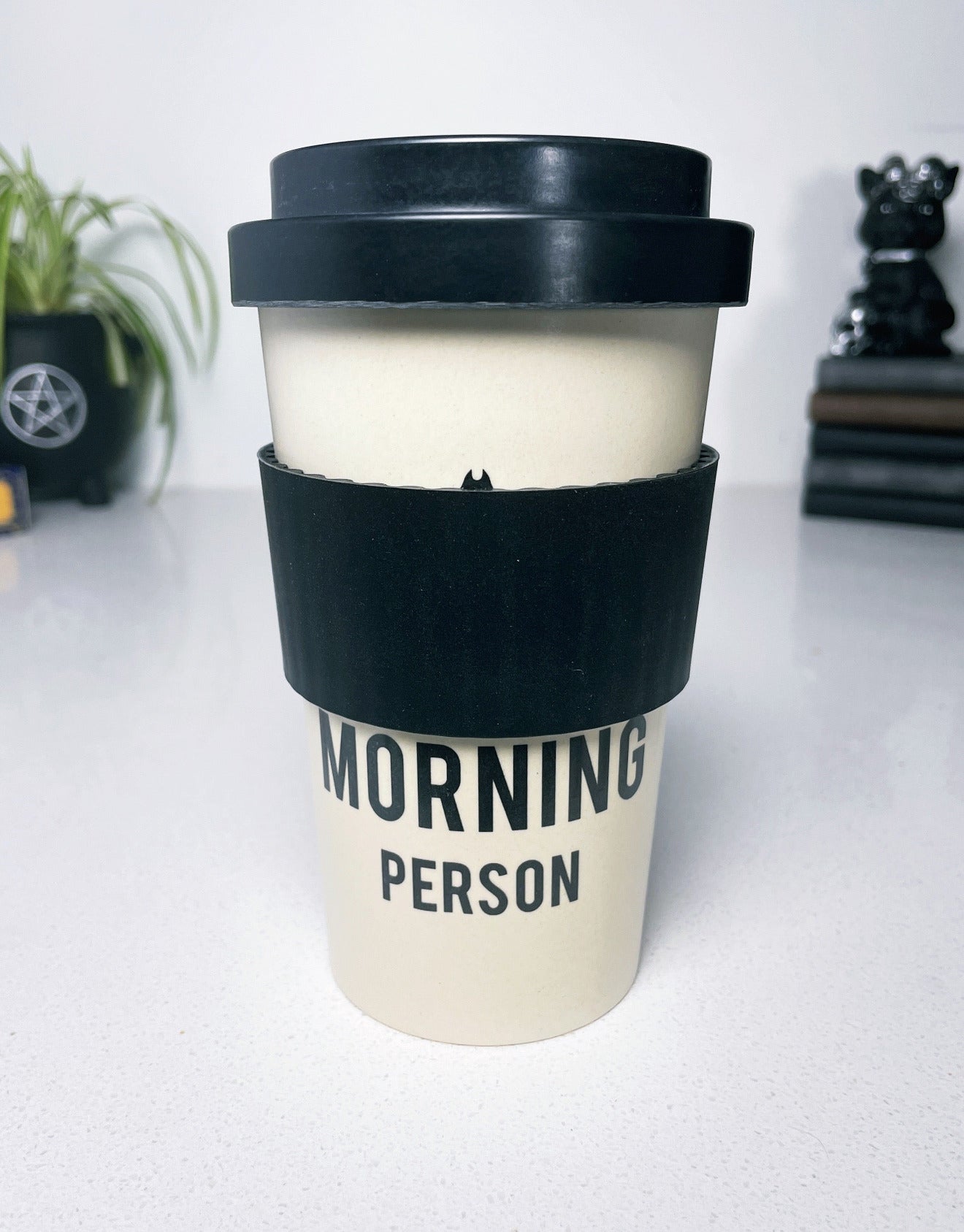 An image of a white travel mug with a black bat design and the phrase "not a morning person" written in bold black letters. The mug is tall and cylindrical with a black plastic lid that has a sipping hole and a sliding tab to open and close it. 