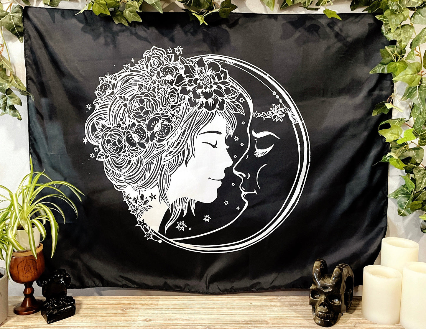 Pictured is a large wall tapestry with a black background and a woman with flowers in her hair smiling at a crescent moon printed in white on it.
