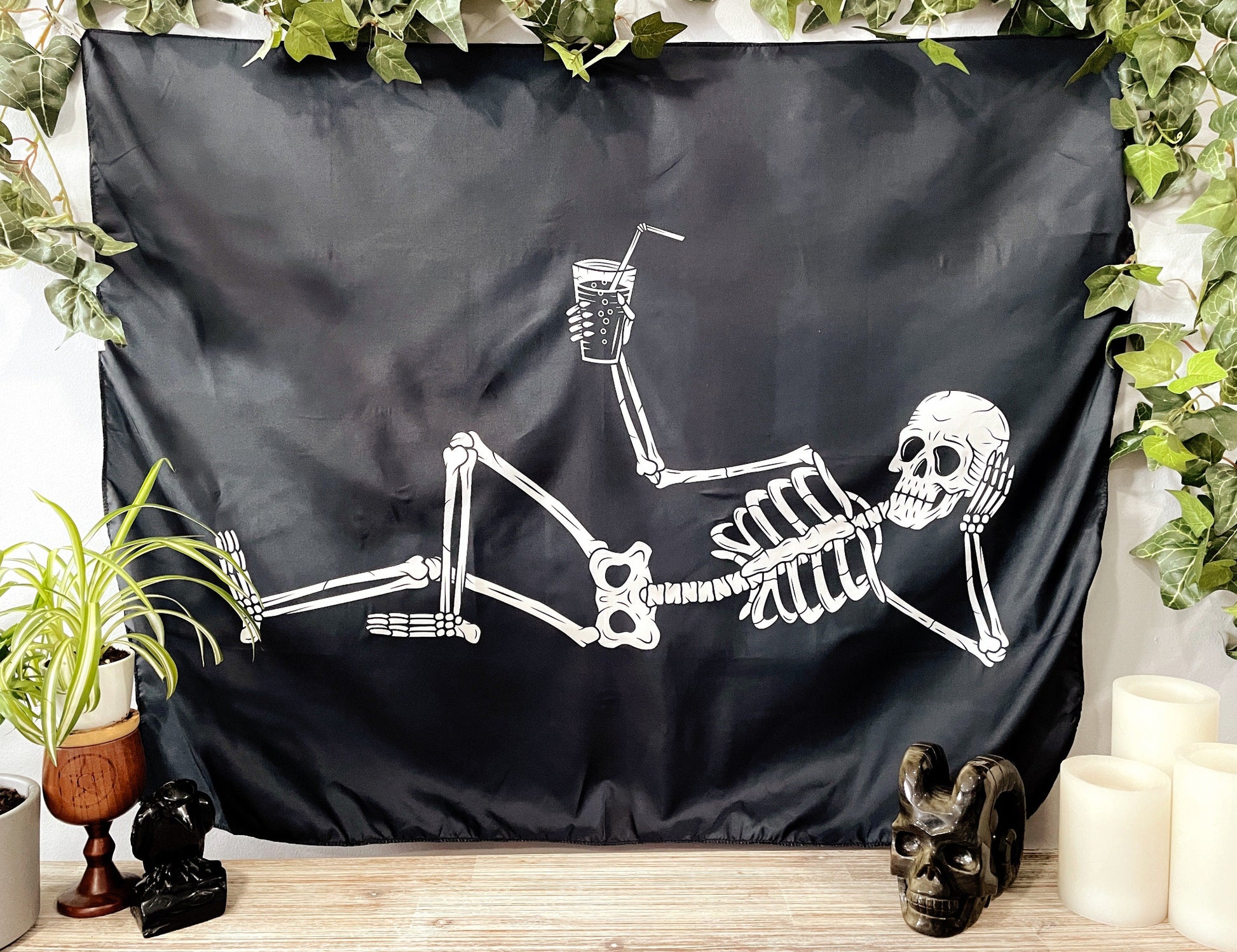 Pictured is a large wall tapestry with a black background and a large skeleton lounging with a drink in its hand printed in white on it.