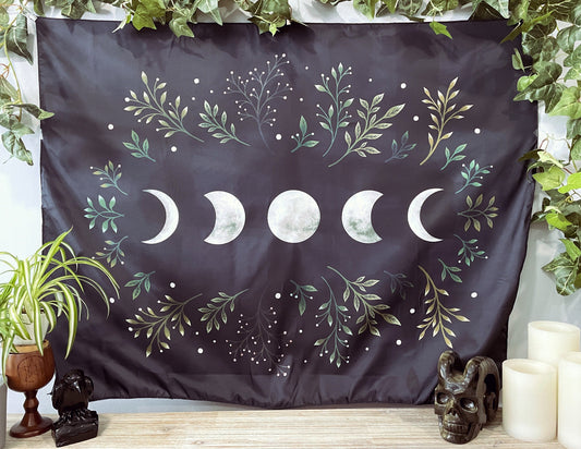 Pictured is a large wall tapestry with a black background and moon phases and leaves printed in white on it.