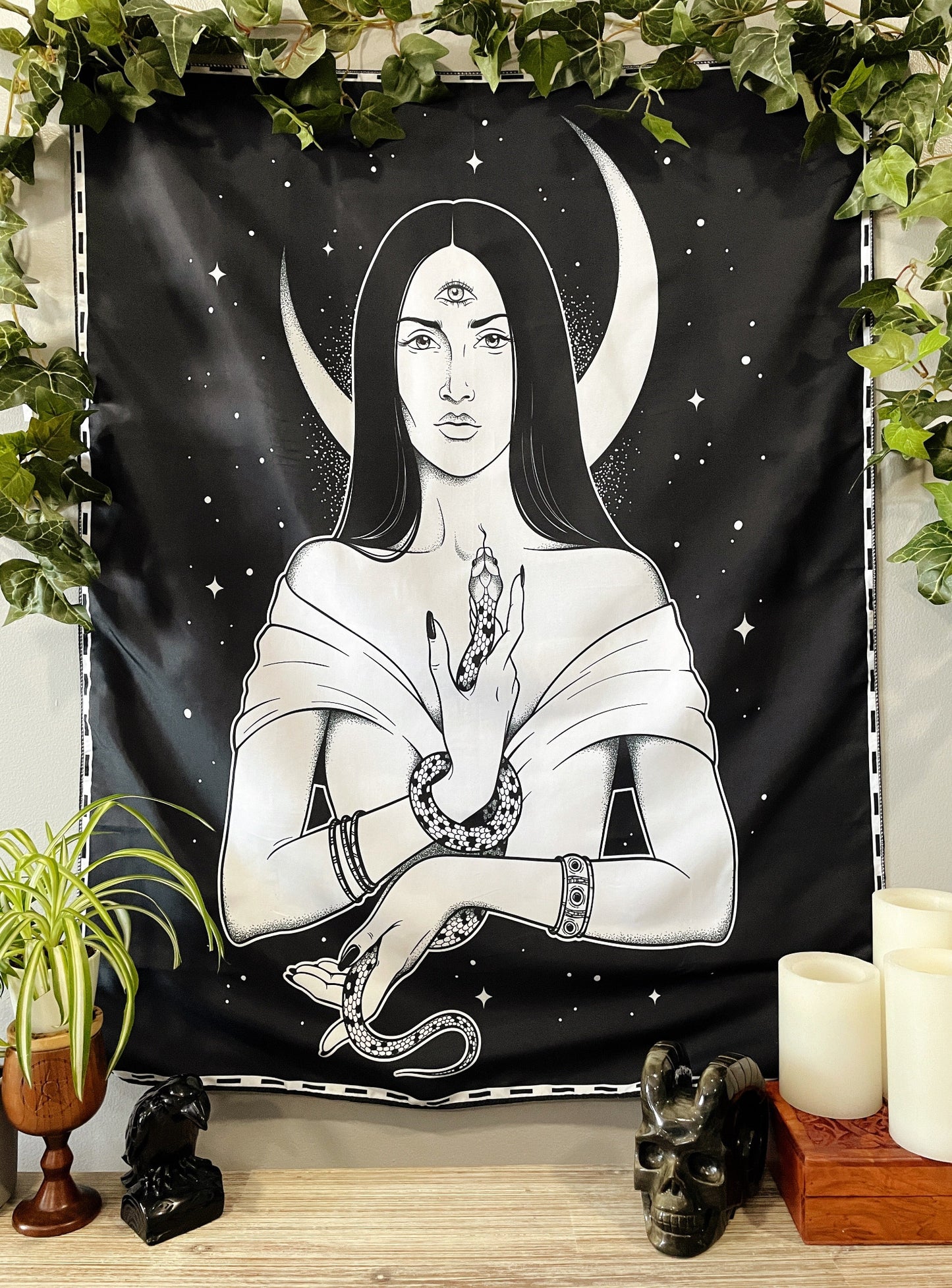 Pictured is a large wall tapestry with a black background and a woman holding a snake with a crescent moon behind her printed in white on it.