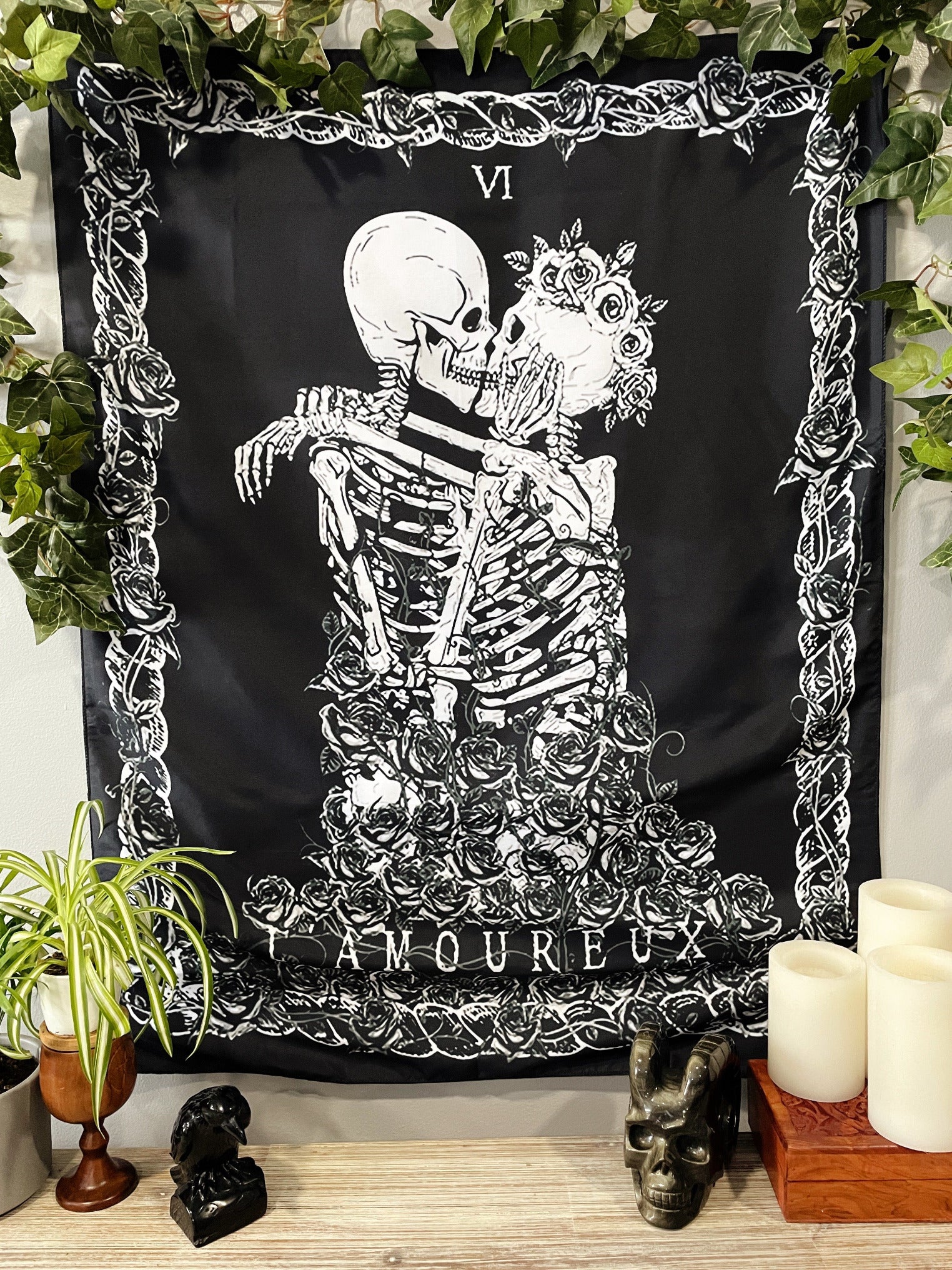 Pictured is a large wall tapestry with a black background and two skeletons kissing on a tarot card "the lovers" printed in white on it.