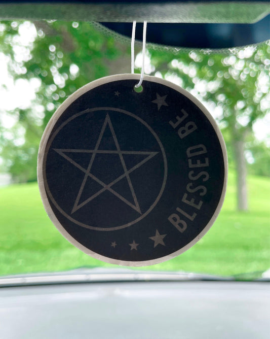 An image of a circular car air freshener featuring a pentagram and a crescent moon on a black background. The words "Blessed Be" are written in white, cursive letters at the bottom of the air freshener.
