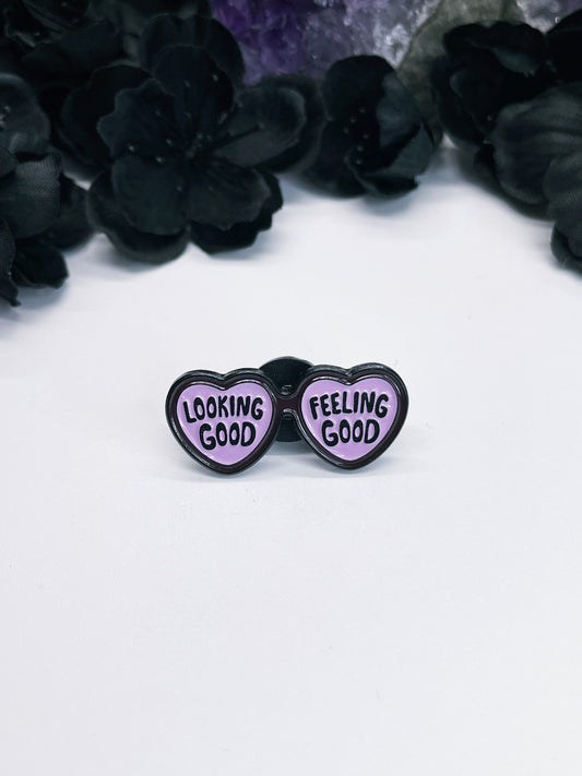 An image of an enamel pin featuring a pair of purple sunglasses with the phrase "Looking good feeling good" written in black letters across the lenses. The purple color of the sunglasses adds a touch of vibrancy and energy to the design, making it a great accessory for any outfit or occasion. 
