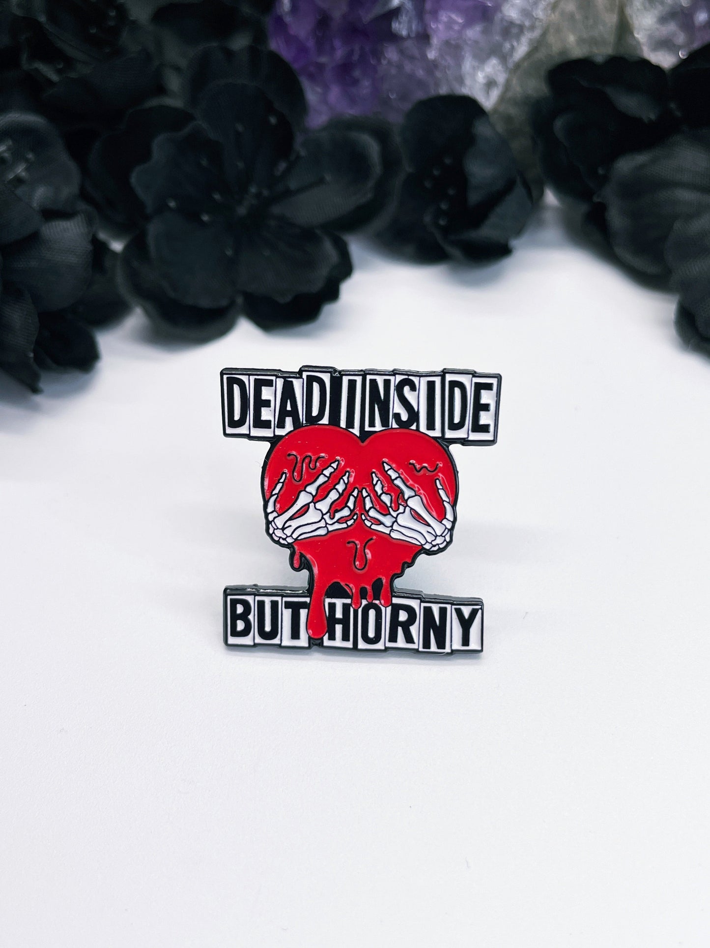 An image of an enamel pin featuring a cartoon-style heart with the words "Dead Inside but Horny" written in bold, white letters across the top.