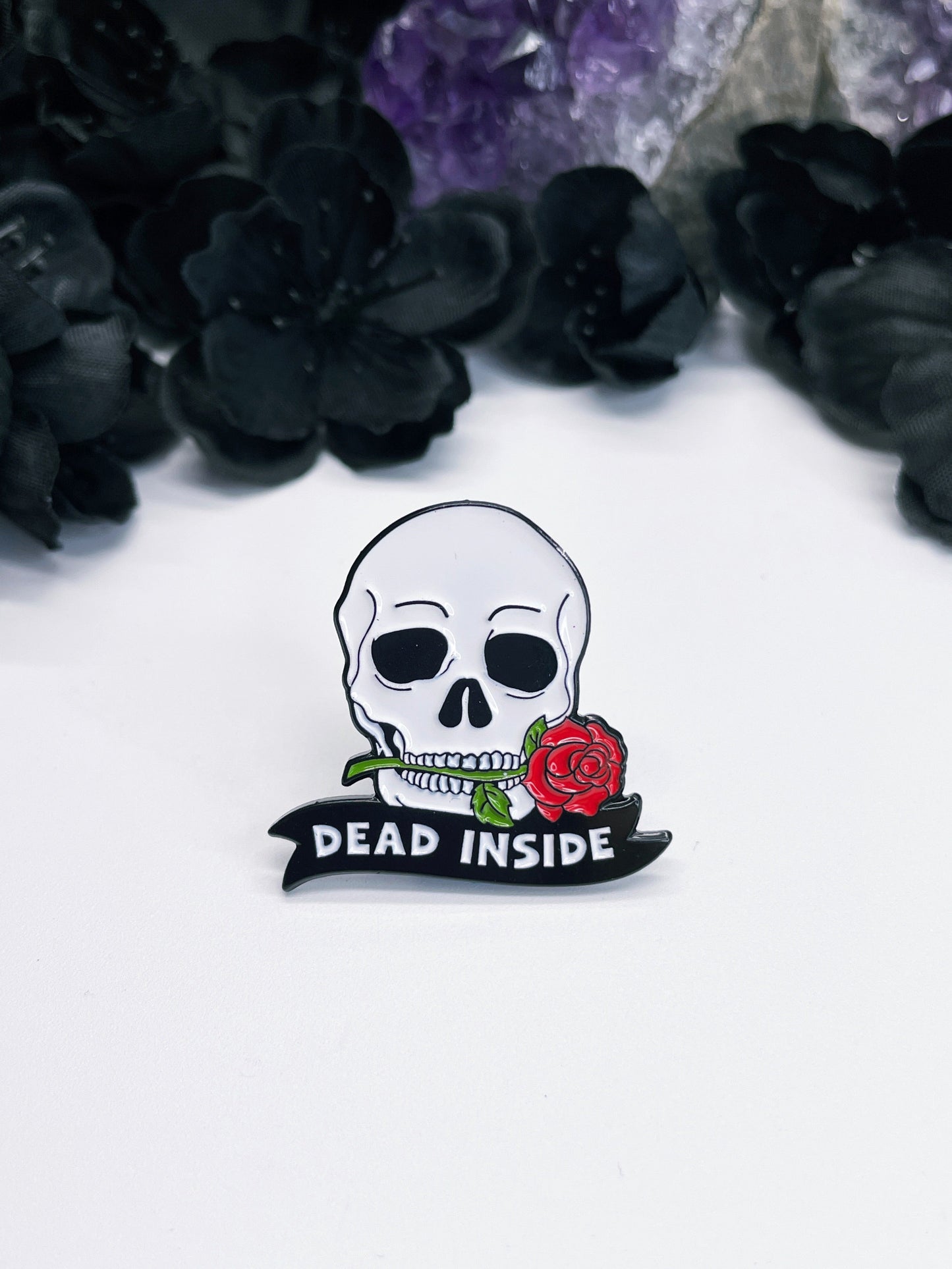 An image of an enamel pin featuring a stylized skeleton skull with a red rose clamped between its teeth, and the words "Dead Inside" written in bold, white letters below it. 