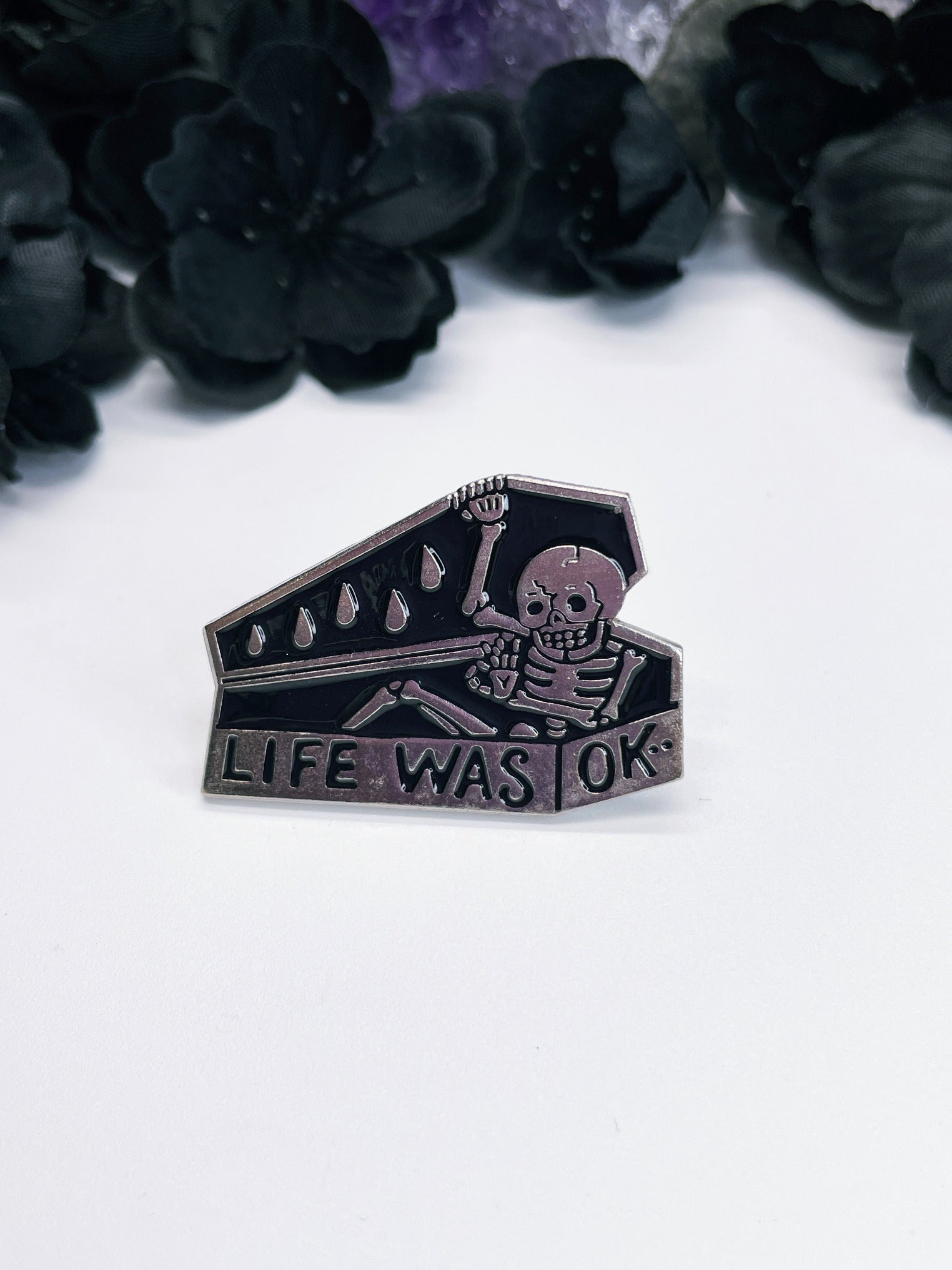 An image of an enamel pin featuring a skeleton emerging from a coffin with the words "Life was okay" written in black letters below it. The pin is coffin-shaped with a silver border and has a metal fastener on the back. The pin is a humorous and ironic take on life and death, and is perfect for anyone who enjoys dark humor or has a gothic and macabre style. 