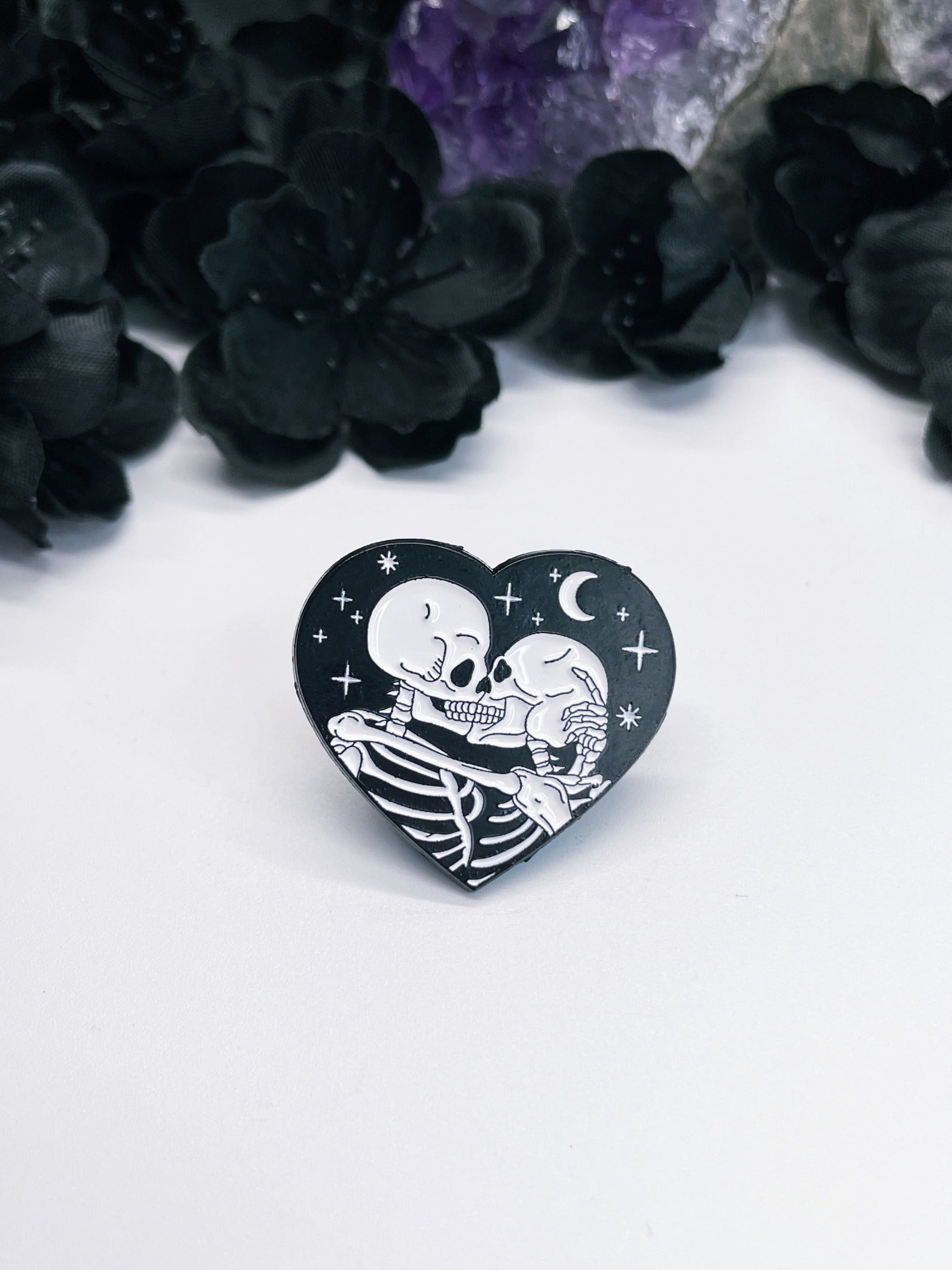 Pictured is an enamel pin of two skeletons kissing inside a heart.