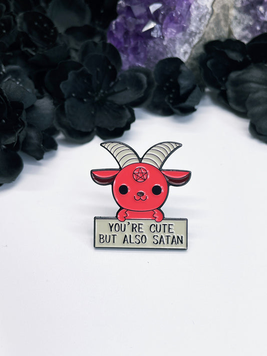 An enamel pin featuring a cute red cartoon of baphomet holding a sign that says "you're cute but also satan" on it.