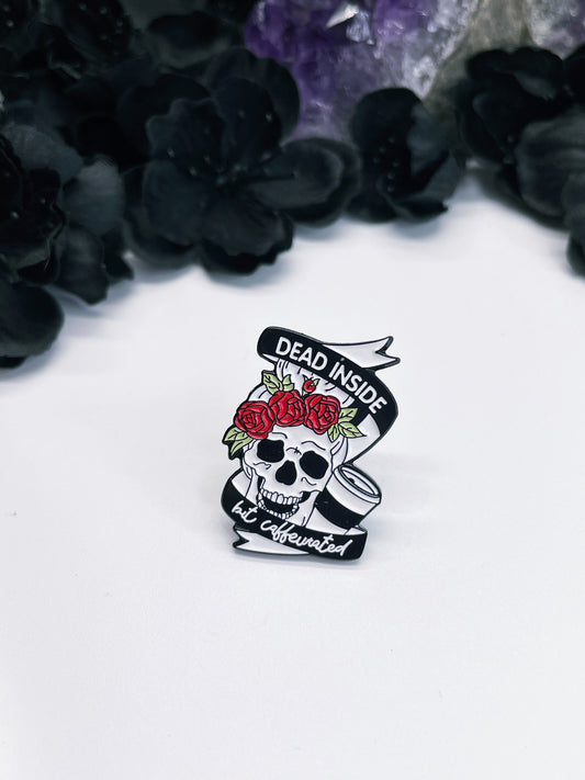 An image of an enamel pin featuring a cartoon-style skull holding a coffee cup. The words "Dead Inside but Caffeinated" are written in bold, white letters above and below the skull. 