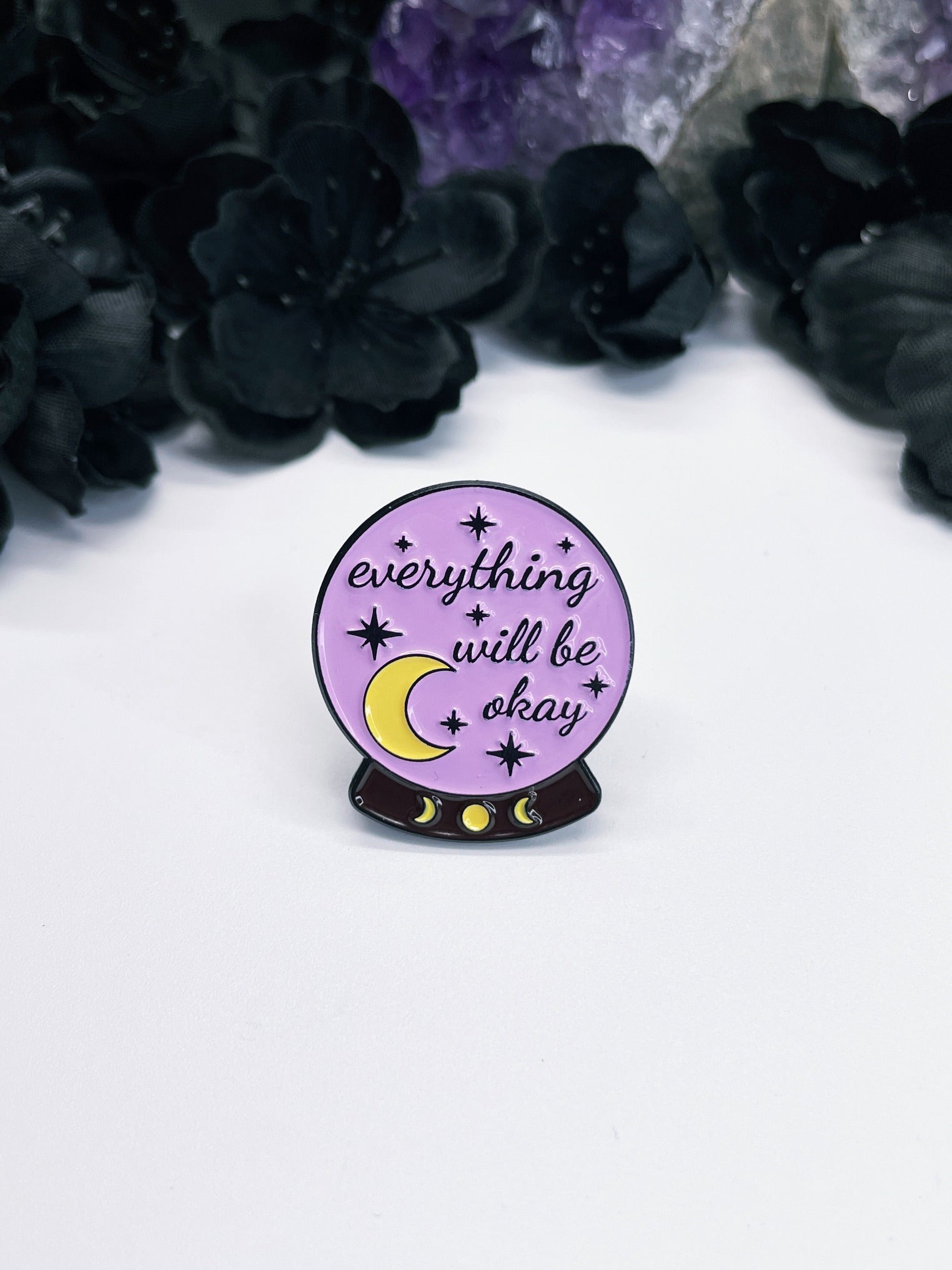 An image of an enamel pin featuring a crystal ball with the words "Everything Will Be Okay" written inside in black lettering. The pin is circular with a black border and has two metal fasteners on the back. 