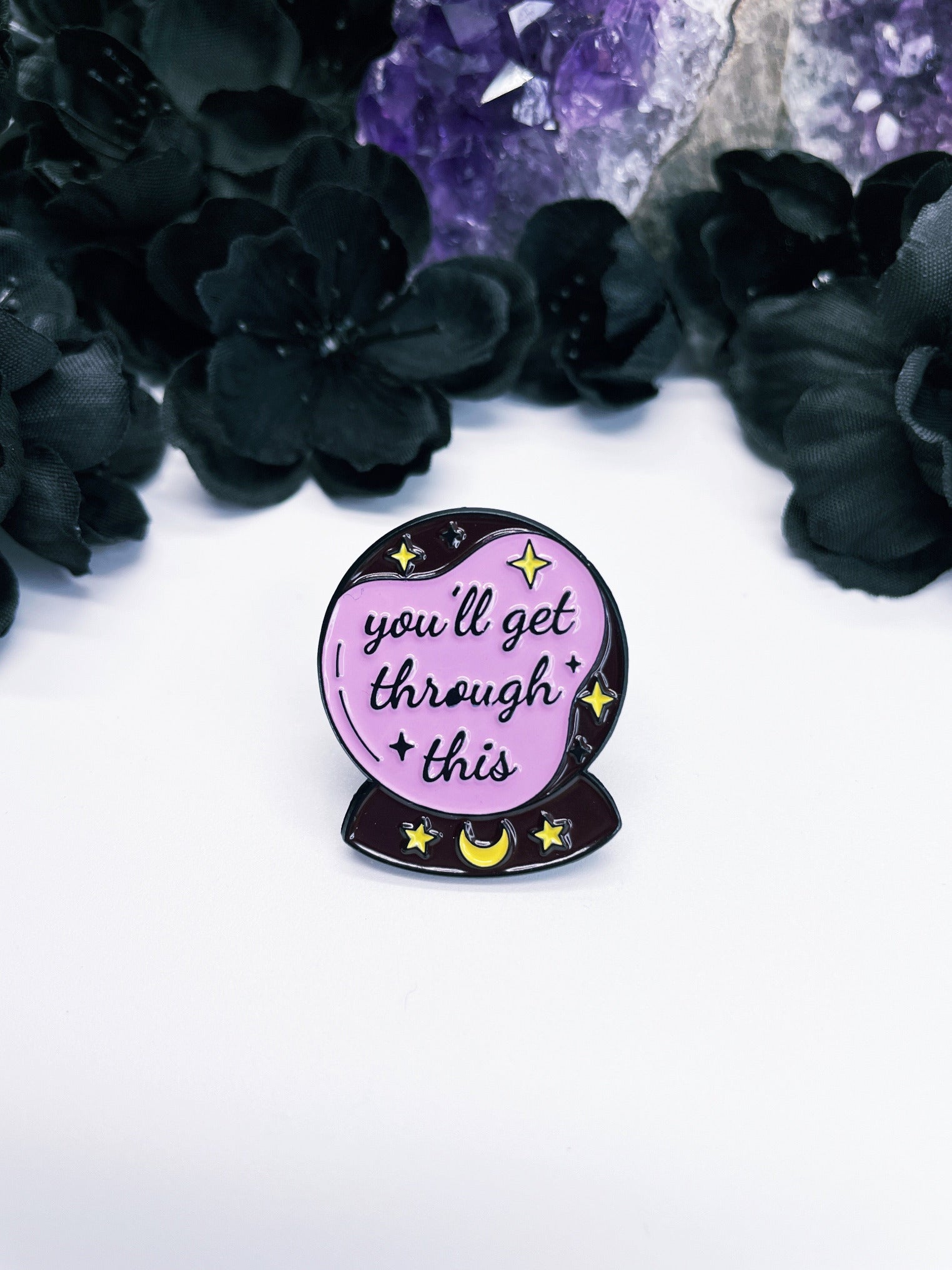 An enamel pin featuring a purple crystal ball with the words "you'll get through this" in black letters.