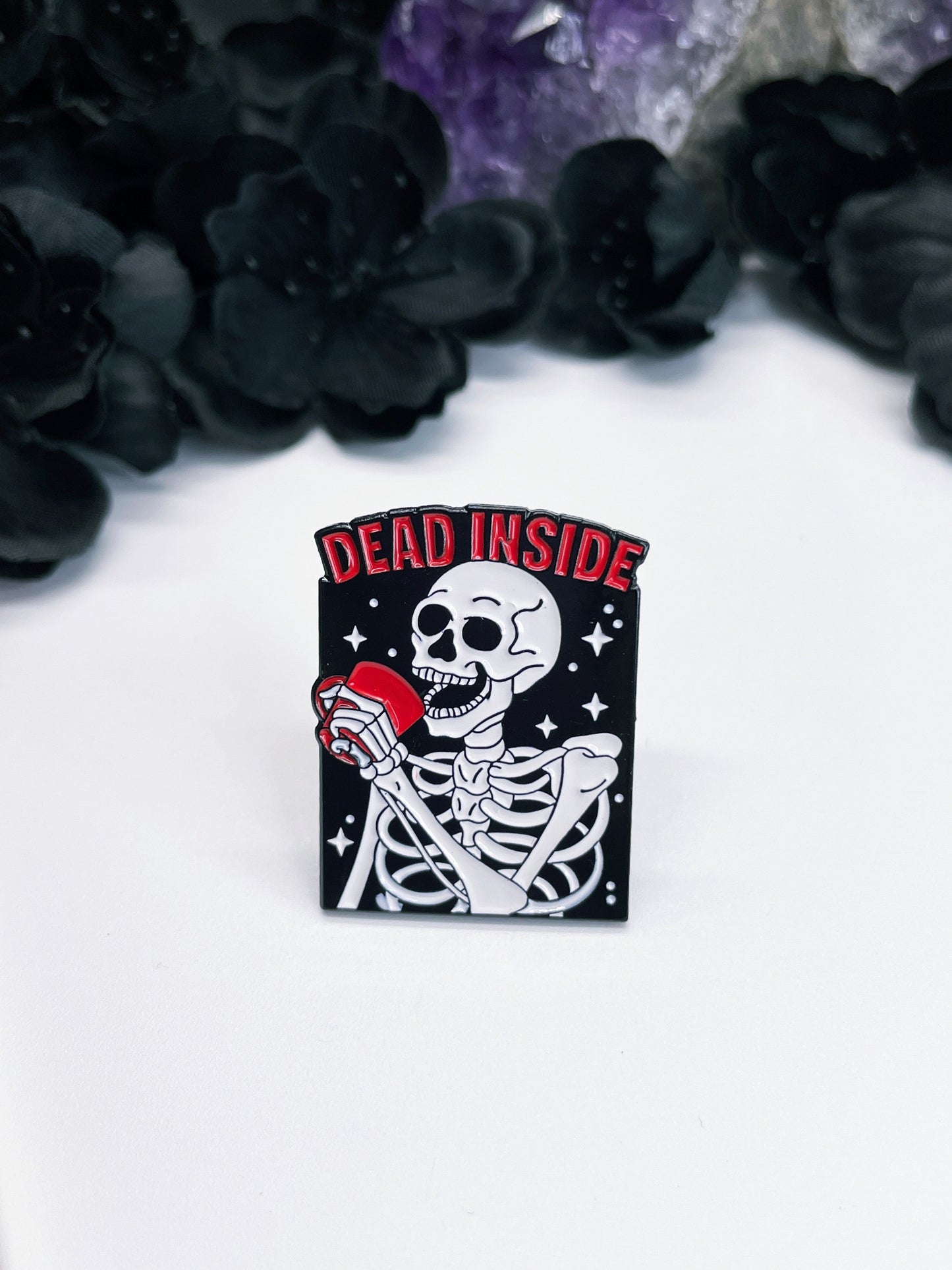 Alt text: An image of an enamel pin featuring a cartoon-style skeleton holding a coffee mug with the words "Dead Inside" written in bold, red letters above it.