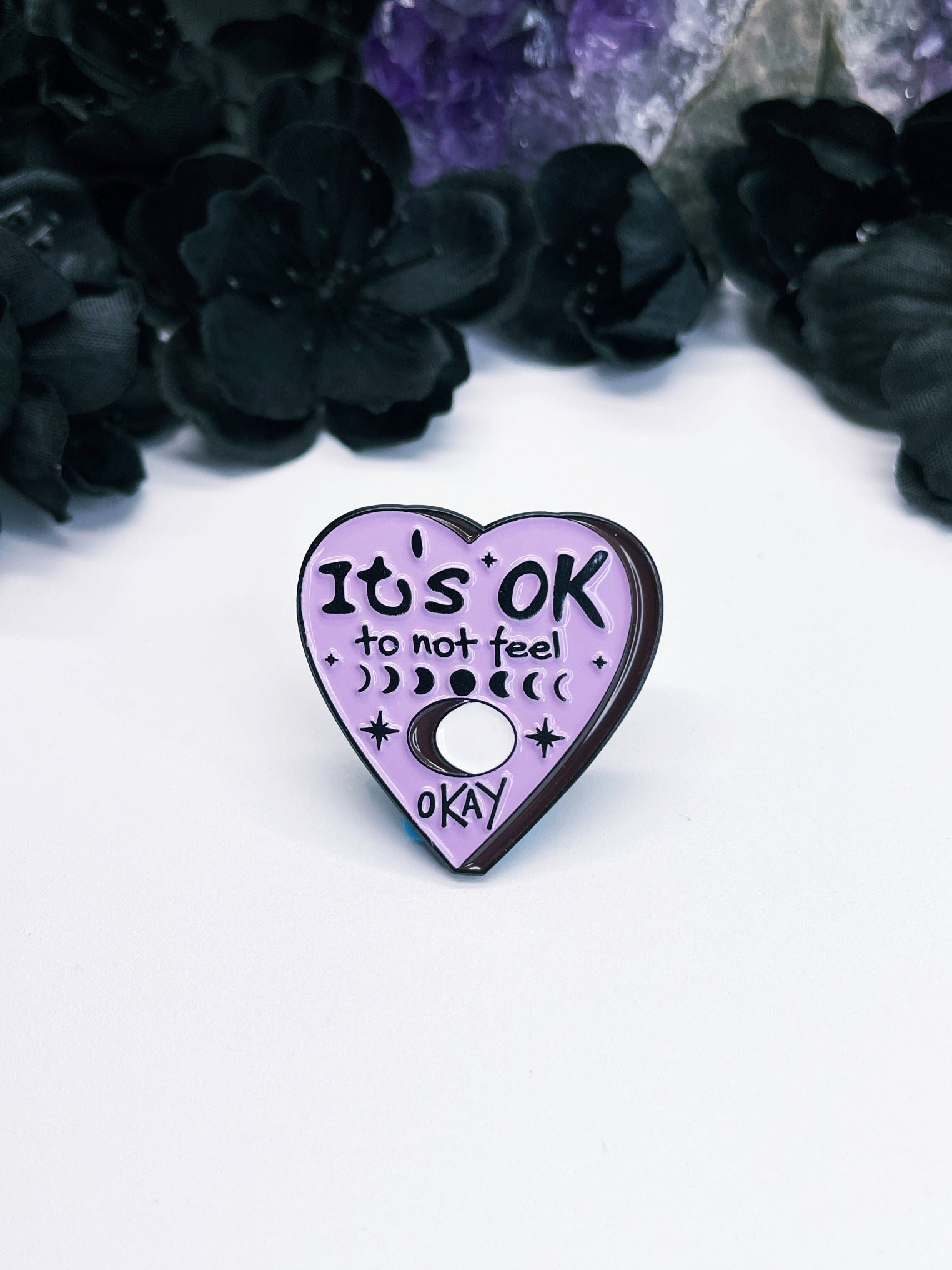 An image of an enamel pin featuring a purple heart with the words "It's Okay to Not Feel Okay" written in black letters. The pin is a simple yet powerful accessory that promotes mental health awareness and encourages people to embrace their emotions and seek help when needed. It is a meaningful and inspiring reminder for anyone who is struggling with their mental health or supporting someone who is going through a tough time.