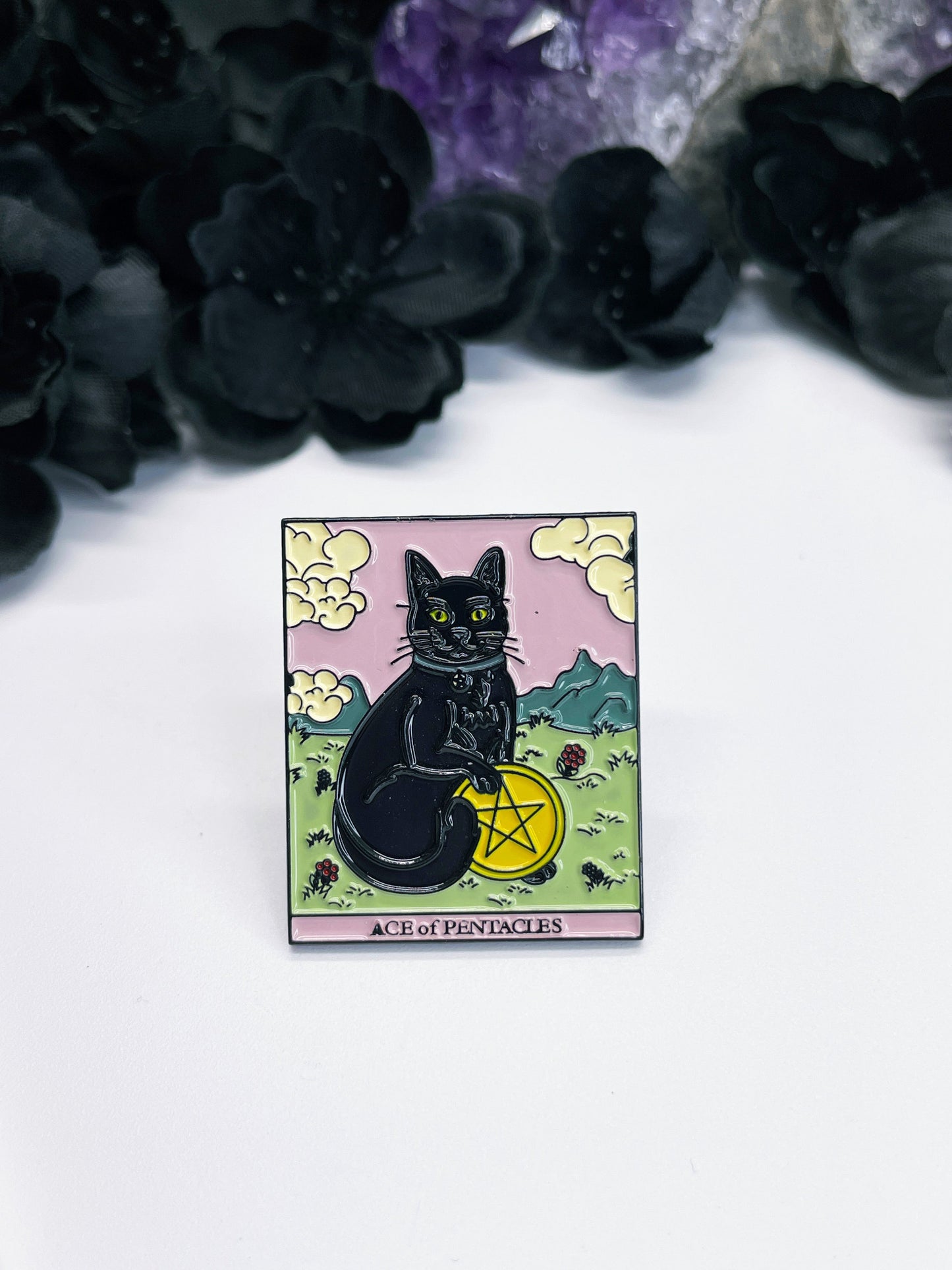 An image of an enamel pin featuring the Ace of Pentacles tarot card with a black cat perched on top of a golden coin. The card is depicted with intricate details and bright colors, including green leaves and pink flowers surrounding the coin. The words "Ace of Pentacles" are written in elegant script at the bottom of the card. The black cat, with its piercing yellow eyes and arched back, adds a touch of mystery and magic to the design. 
