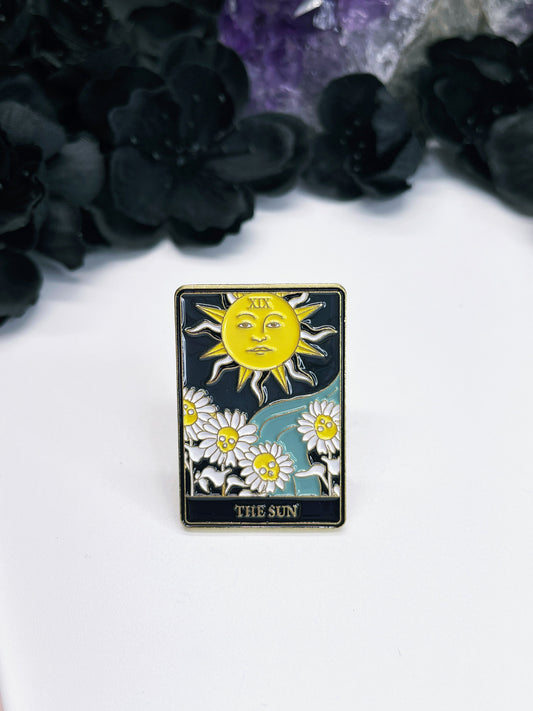  An enamel pin featuring a tarot card with a yellow sun and flowers on it, the card is "The Sun."