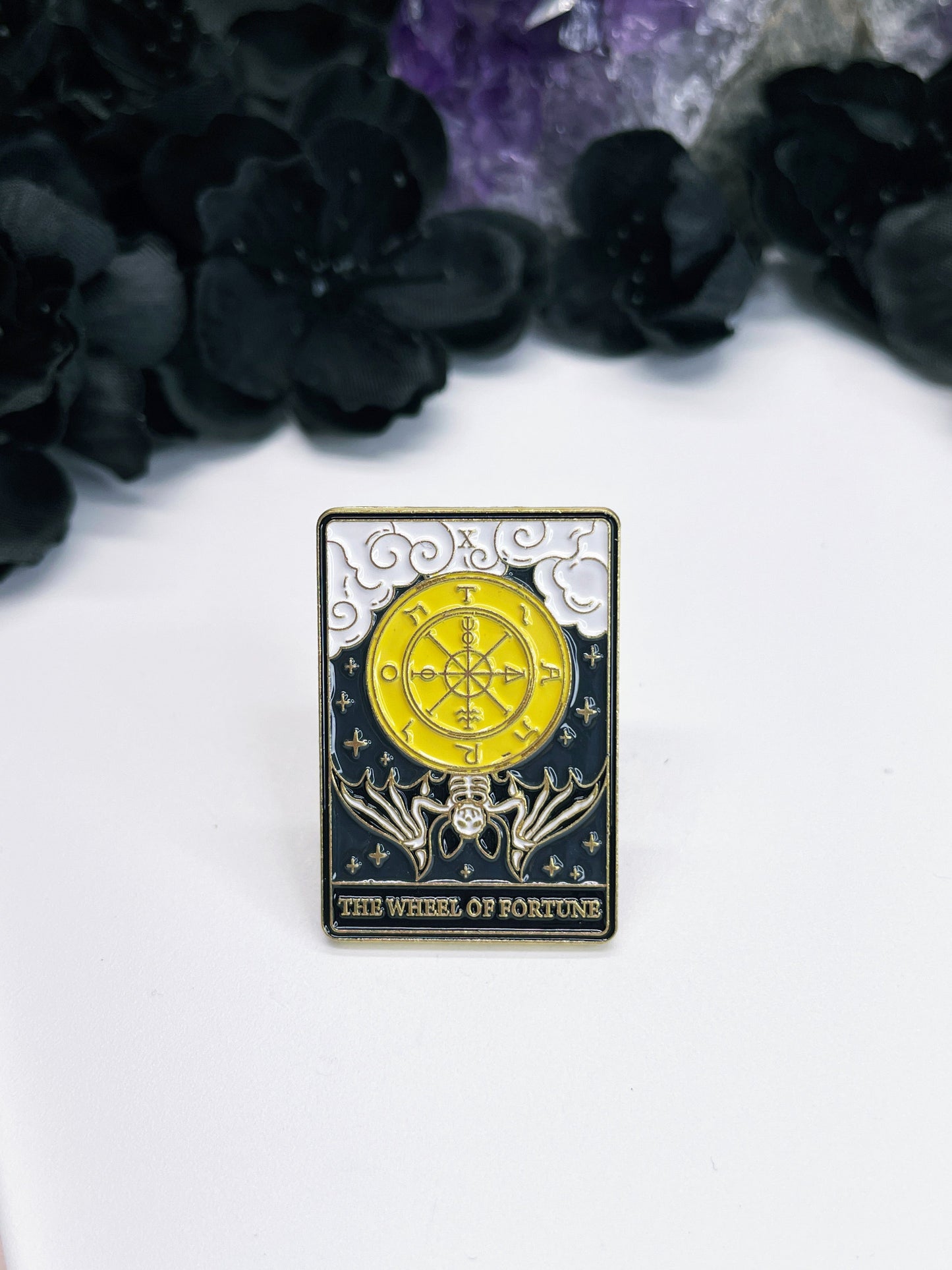  An enamel pin featuring a tarot card with a wheel and a bat on it, the card is "The Wheel of Fortune."