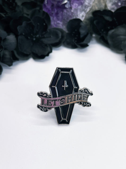An image of an enamel pin featuring a black coffin with the words "Let's Hide" written in black letters on the top. The pin is a fun and quirky accessory that celebrates the spooky and gothic aesthetic, and is perfect for anyone who loves Halloween or has a dark and edgy style. 