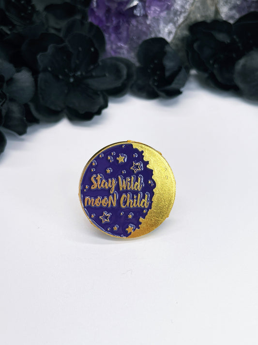 An enamel pin featuring a purple and gold moon with the words "stay wild moon child" written on it in gold letters. The pin is on a white background.