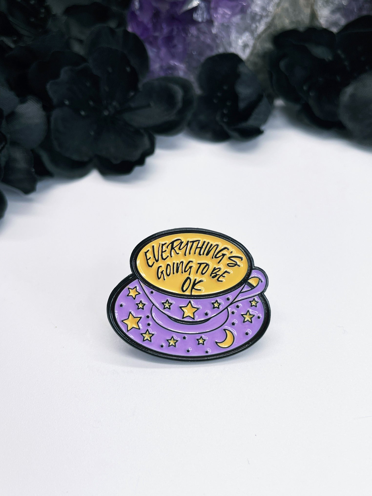 An image of an enamel pin featuring a teacup with the words "Everything's Going To Be Okay" written inside in black lettering. The pin has a black border and has metal fasteners on the back. 
