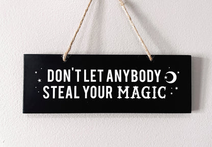 An image of a black sign with white letters that reads "Don't Let Anybody Steal Your Magic." The sign is rectangular and has a thick rope hanger attached to it, making it suitable for hanging on a wall or door. 