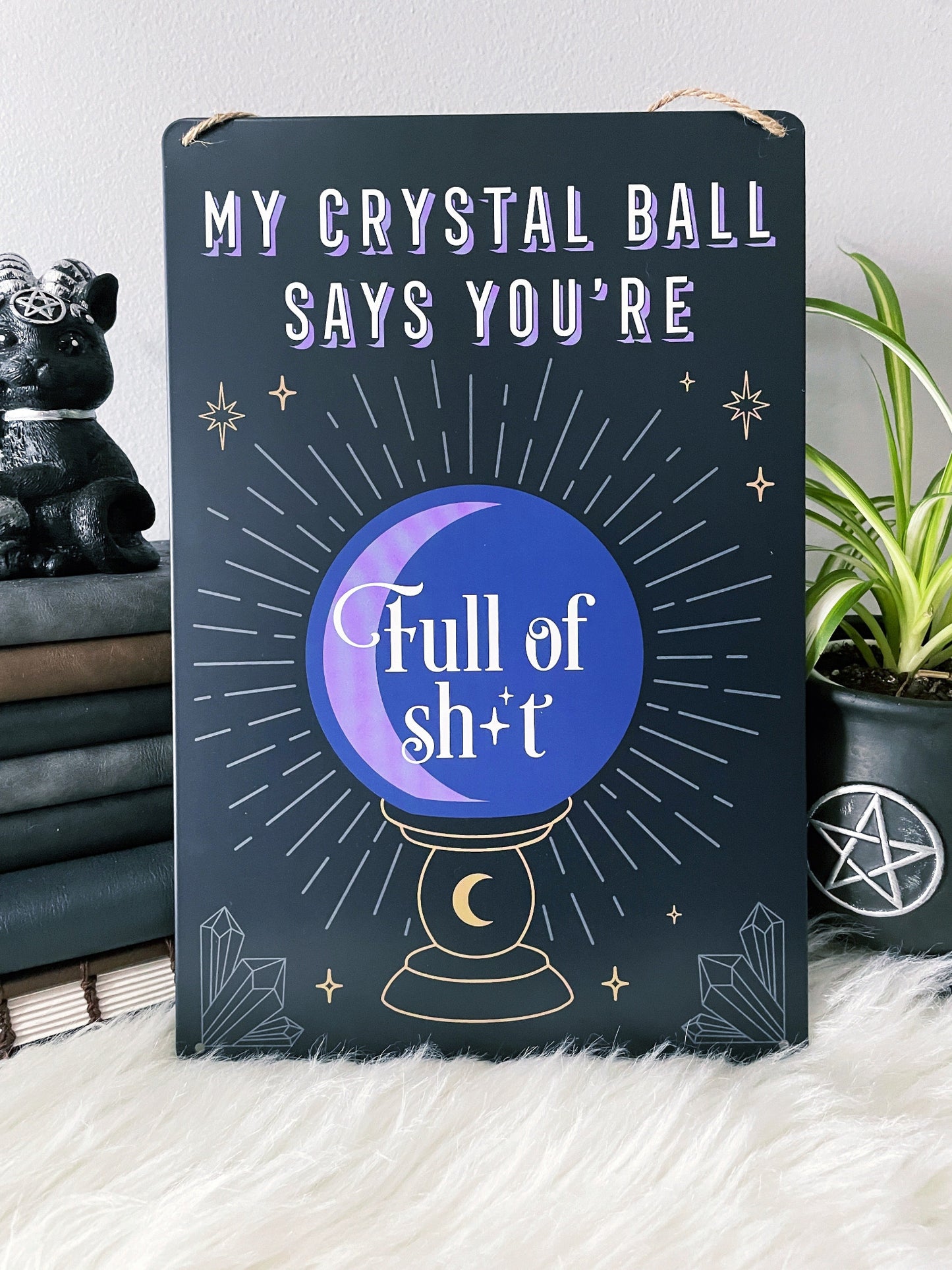 An image of a metal wall sign featuring a crystal ball design with the phrase "My crystal ball says you're full of shit" written in bold letters. The crystal ball design is illustrated in white against a deep purple background, with intricate lines and details that create a mystical and magical look.
