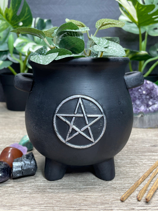 Pictured is a black terracotta plant holder in the shape of a cauldron with a pentagram on it.