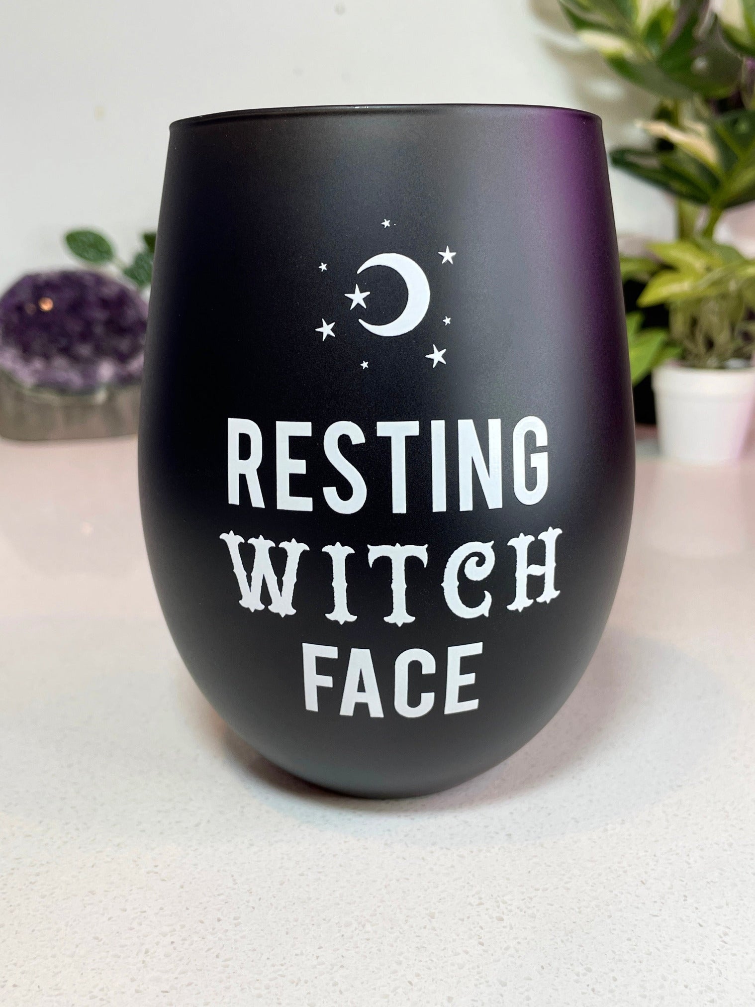 A black stemless wine glass with the words "resting witch face" written on it in white letters. The glass is on a white background.