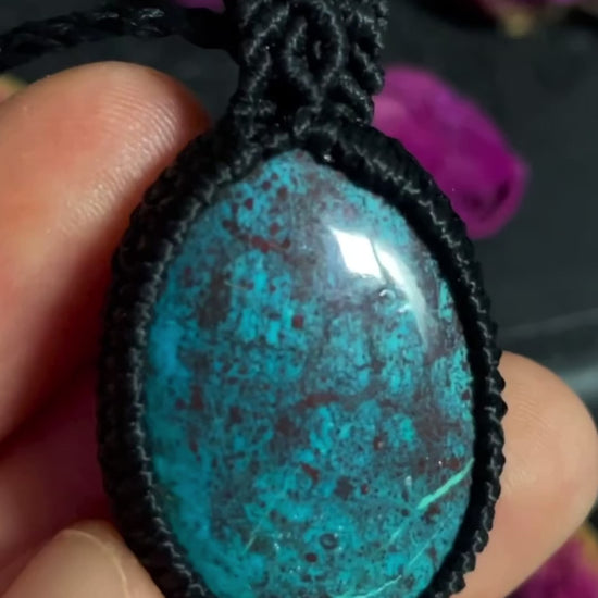 Pictured is a chrysocolla shattuckite and cuprite cabochon wrapped in macrame thread. A gothic book and flowers are nearby.