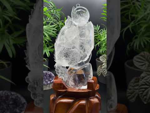 Pictured is a gorgeous clear quartz carving of a dragon holding a crystal wand and chasing a lucky sphere. It stands on a custom, hand-carved wood stand.